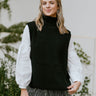 Front view of model wearing the Brooklyn Sweater Vest in Black, that features black knit fabric, a turtleneck neckline, a high- low hem with side slits, and a sleeveless body