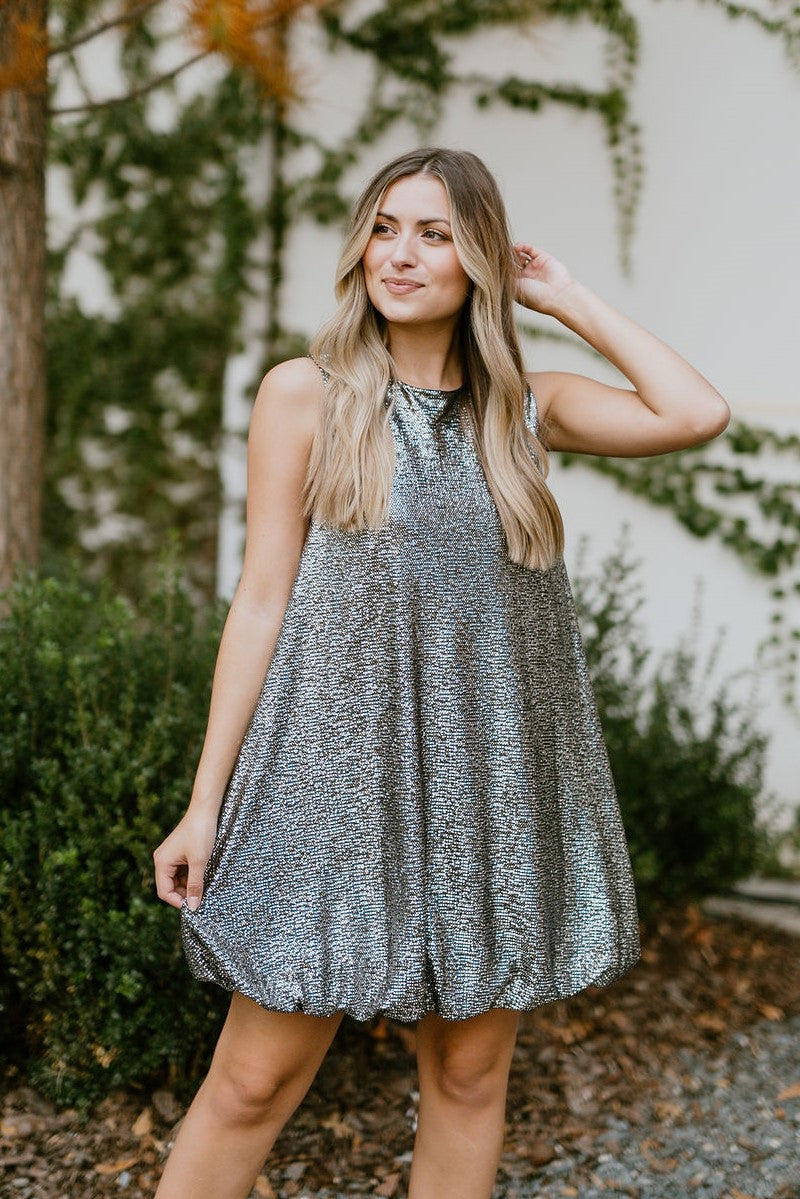 Front view of model wearing the Let's Celebrate Dress that has black fabric with silver speckles, a round neckline, a back button closure, a bubble hem, and a sleeveless design.