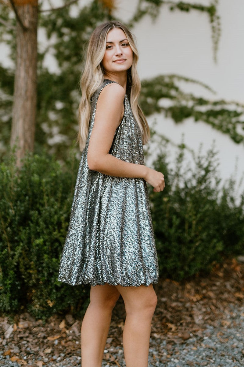 Side view of model wearing the Let's Celebrate Dress that has black fabric with silver speckles, a round neckline, a back button closure, a bubble hem, and a sleeveless design.