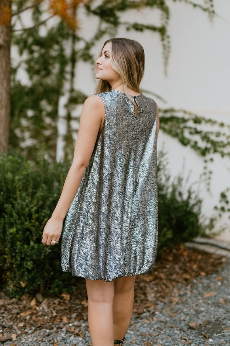 Back view of model wearing the Let's Celebrate Dress that has black fabric with silver speckles, a round neckline, a back button closure, a bubble hem, and a sleeveless design.