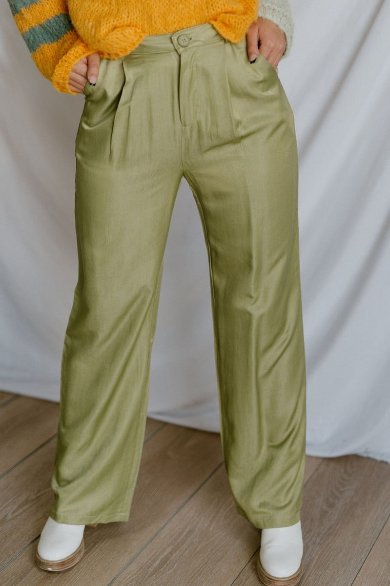 Front view of model wearing the Real Deal Satin Pants in Green that have light green satin fabric, two front pockets, two back pockets, a front zipper and button closure, belt loops, pleated detailing, and wide legs