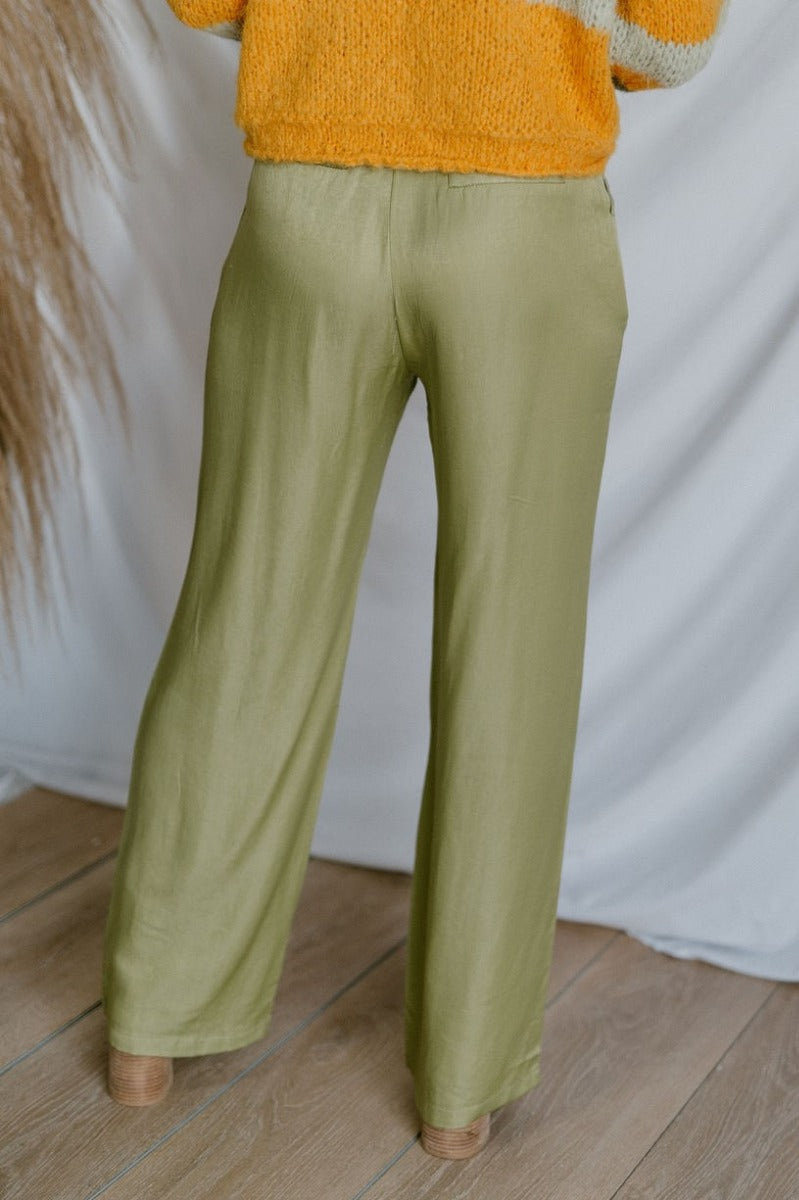 Back view of model wearing the Real Deal Satin Pants in Green that have light green satin fabric, two front pockets, two back pockets, a front zipper and button closure, belt loops, pleated detailing, and wide legs