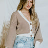 Frontal side view of model wearing the You're A Keeper Cropped Cardigan that has knit fabric with a cream and brown chevron pattern, cream ribbed trim, tortoise buttons, a v neck, a cropped waist, and long balloon sleeves.