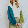 Front view of model wearing the Pop Of Color Sweater that has cream cable knit fabric, teal fabric on the right arm, lime fabric on the left arm, a turtleneck, a cropped waist, and long sleeves