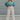 Full body front view of model wearing the It Girl Pants that have natural color fabric, pleated details, two front pockets, a front zipper and hook closure, an elastic-back waistband with belt loops, and flare pant legs.