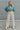 Full body front view of model wearing the It Girl Pants that have natural color fabric, pleated details, two front pockets, a front zipper and hook closure, an elastic-back waistband with belt loops, and flare pant legs.