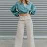 Full body front view of model wearing the It Girl Pants that have natural color fabric, pleated details, two front pockets, a front zipper and hook closure, an elastic-back waistband with belt loops, and flare pant legs.