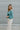 Clsoe side view of model wearing the It Girl Pants that have natural color fabric, pleated details, two front pockets, a front zipper and hook closure, an elastic-back waistband with belt loops, and flare pant legs.