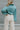Close back view of model wearing the It Girl Pants that have natural color fabric, pleated details, two front pockets, a front zipper and hook closure, an elastic-back waistband with belt loops, and flare pant legs.