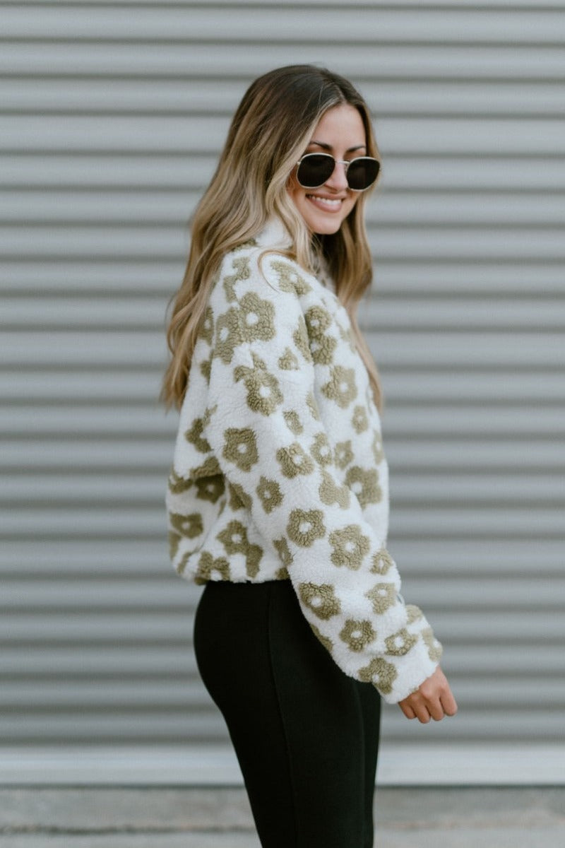 Side view of model wearing The In Bloom Teddy Jacket that has white teddy fabric with an olive flower pattern, a quarter zip closure with a collar, a cropped waist with a drawstring hem, and long sleeves.