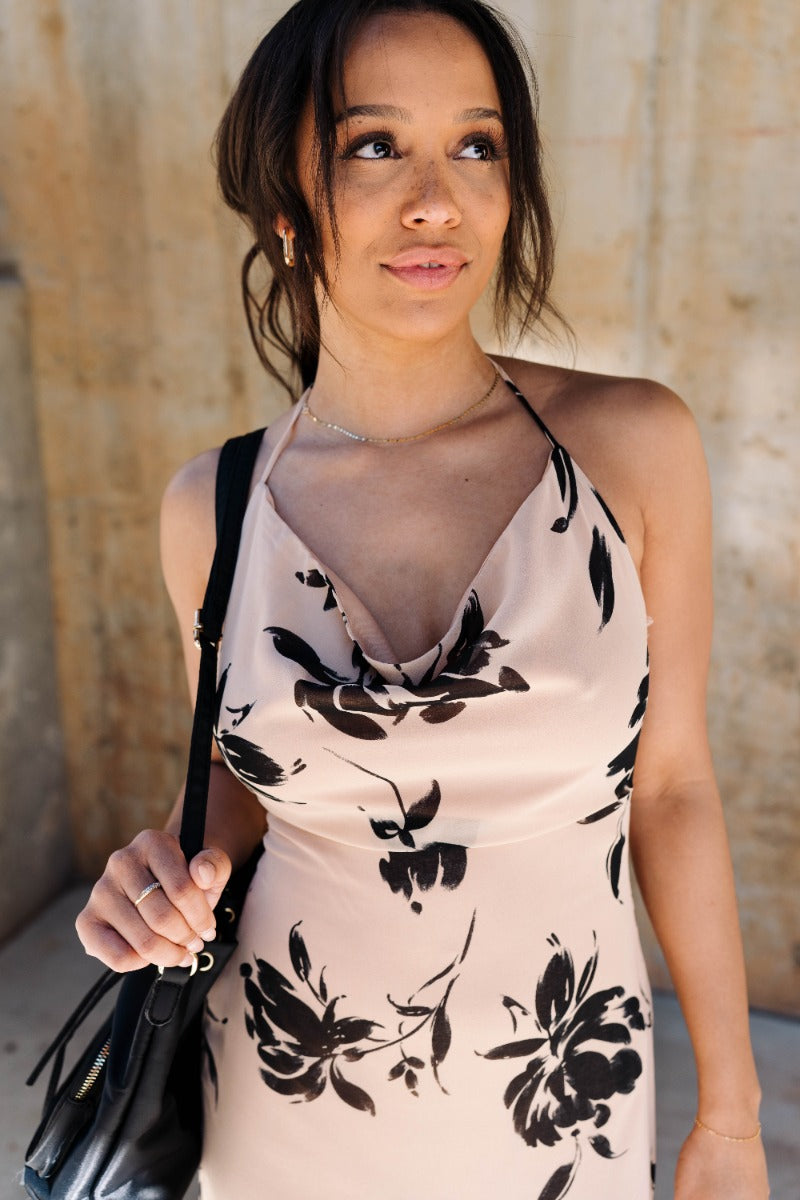 Upper front view of model wearing the Felicity Beige & Black Floral Halter Midi Dress that has khaki sheer fabric with a black floral print, a slit on the side, a halter neck, an open back and a back zipper with a hook closure.