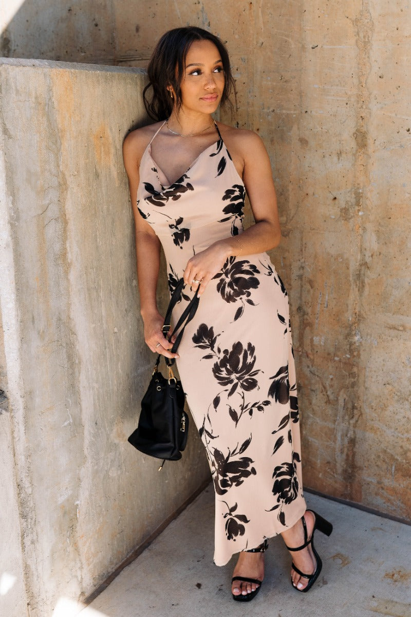 Full body front view of model wearing the Felicity Beige & Black Floral Halter Midi Dress that has khaki sheer fabric with a black floral print, a slit on the side, a halter neck, an open back and a back zipper with a hook closure.