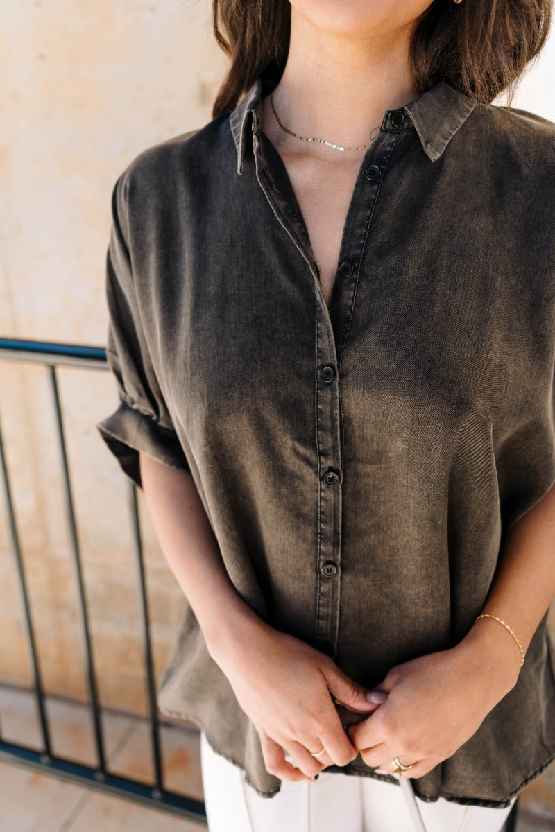 Close up front view of model wearing the Grace Washed Black Short Sleeve Top that has light washed black tencel fabric, monochrome front button up closures, a collared neckline and short sleeves.
