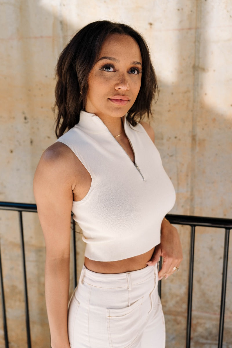 Frontal side view of model wearing the Nova Cream Knit Quarter Zip Up Tank which features cream knit fabric, cropped waist, high neckline, quarter zip up closure and sleeveless.