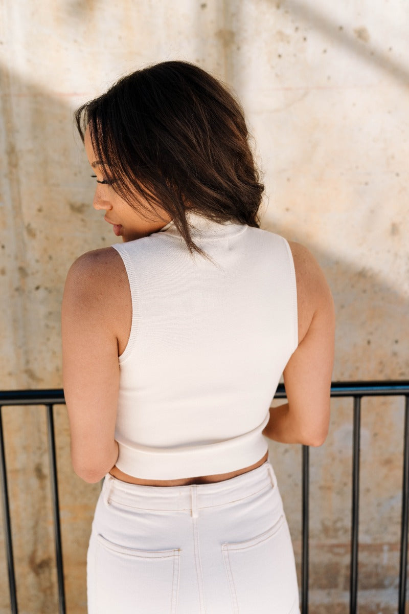 Back view of model wearing the Nova Cream Knit Quarter Zip Up Tank which features cream knit fabric, cropped waist, high neckline, quarter zip up closure and sleeveless.