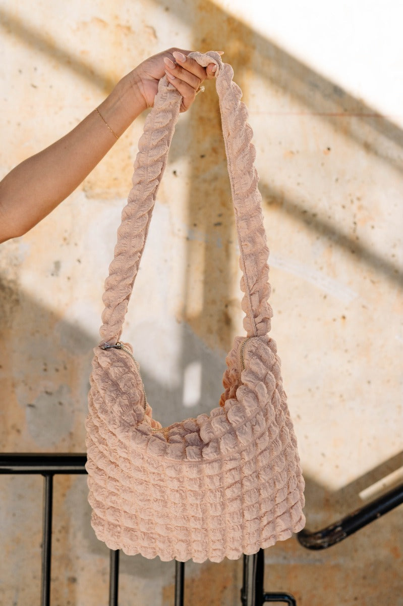 Front view of model holding the Ashley Bubble Tote Bag in Sand which features sand textured bubble fabric, one shoulder strap, lavender lining and monochrome zipper closure with silver hardware.