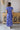 Back view of model wearing the Summertime Blues Maxi Dress that features royal blue fabric with a white floral print, maxi length, a slit on the side, a front tie closure, a surplice neckline and short sleeves
