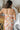 Upper back view of model wearing the Your Truly Floral Midi Dress that has light pink fabric with a coral, yellow, blue, grey and green floral print, midi length, beige lining, a cowl neckline, and tie spaghetti straps