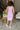 Full body back view of model wearing the Out And About Dress In Lavender which features lavender knit fabric, ruched side details, midi length, lavender lining, a round neckline and a sleeveless design.
