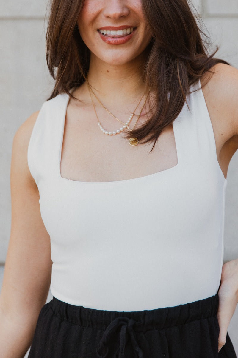 Close up view of model wearing the Luna Cream Sleeveless Bodysuit which features white knit fabric, a square neckline with a racerback, a sleeveless body, and a thong bottom with snap closures.