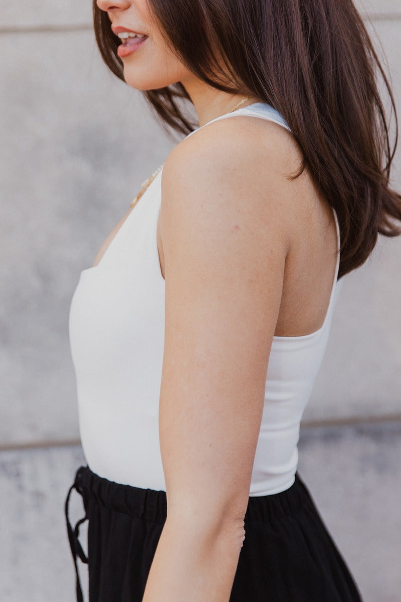 Close up side view of model wearing the Luna Cream Sleeveless Bodysuit which features white knit fabric, a square neckline with a racerback, a sleeveless body, and a thong bottom with snap closures.