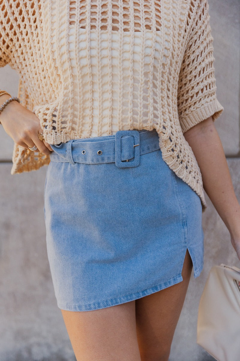 Close up view of model wearing the Daphne Light Washed Denim Belted Skirt which features washed denim fabric, mini length, a small slit detail, a monochrome side zipper with a hook closure, and a monochrome adjustable belt with a buckle.