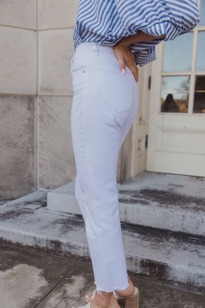 Side view of model wearing the Rebecca White Denim Fray Straight Leg Jeans which features white denim fabric, front zipper with button closure, two front pockets, two back pockets, belt loops, high rise and straight pant legs with fray hem.