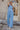 Full body view of model wearing the Everly Chambray Blue Halter Neckline Jumpsuit which features light blue lightweight fabric, pleated details, two front slit pockets, wide pant legs, light blue shorts lining, a halter neckline with a twist detail, a sle
