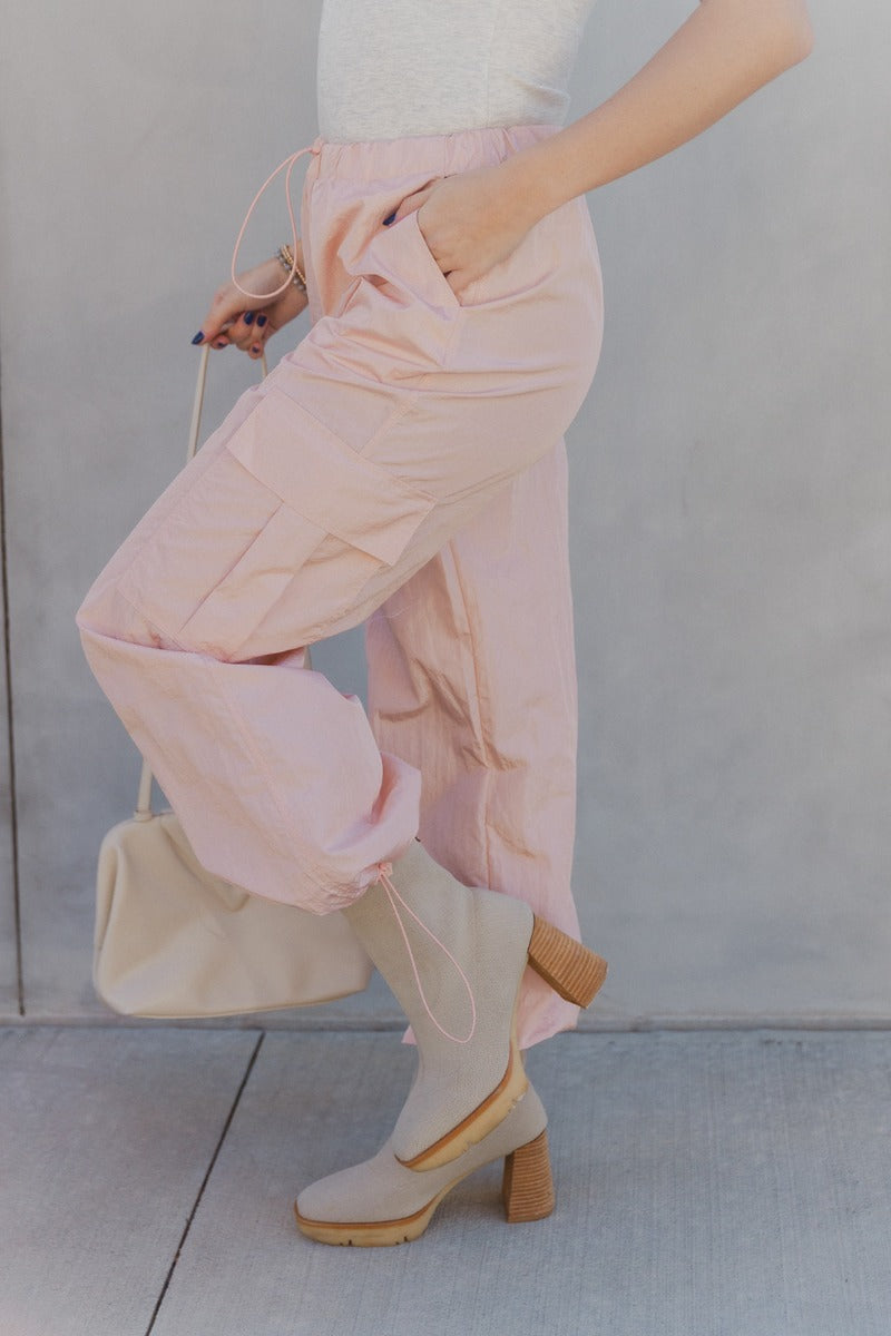 Side view of model wearing the Harper Blush Cargo Drawstring Pants which features blush nylon fabric, two front slit pockets, two side cargo pockets, elastic waistband with drawstring and wide pant legs with drawstring at ankles.
