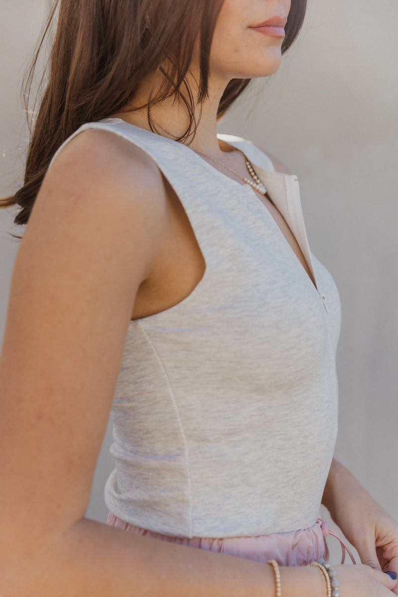 Side view of model wearing the Isla Heather Grey Zip Up Sleeveless Bodysuit which features light heather grey knit fabric, a monochrome front zip up, a round neckline, a sleeveless body, and a thong bottom with snap closures.