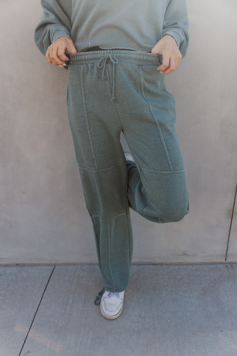 Front view of model wearing the Mia Grey Green Knit Pants which features gray green knit fabric, ribbed hem details, two front pockets, one back pockets, elastic waistband with drawstring ties and wide pant legs with drawstring ties at the ankles.
