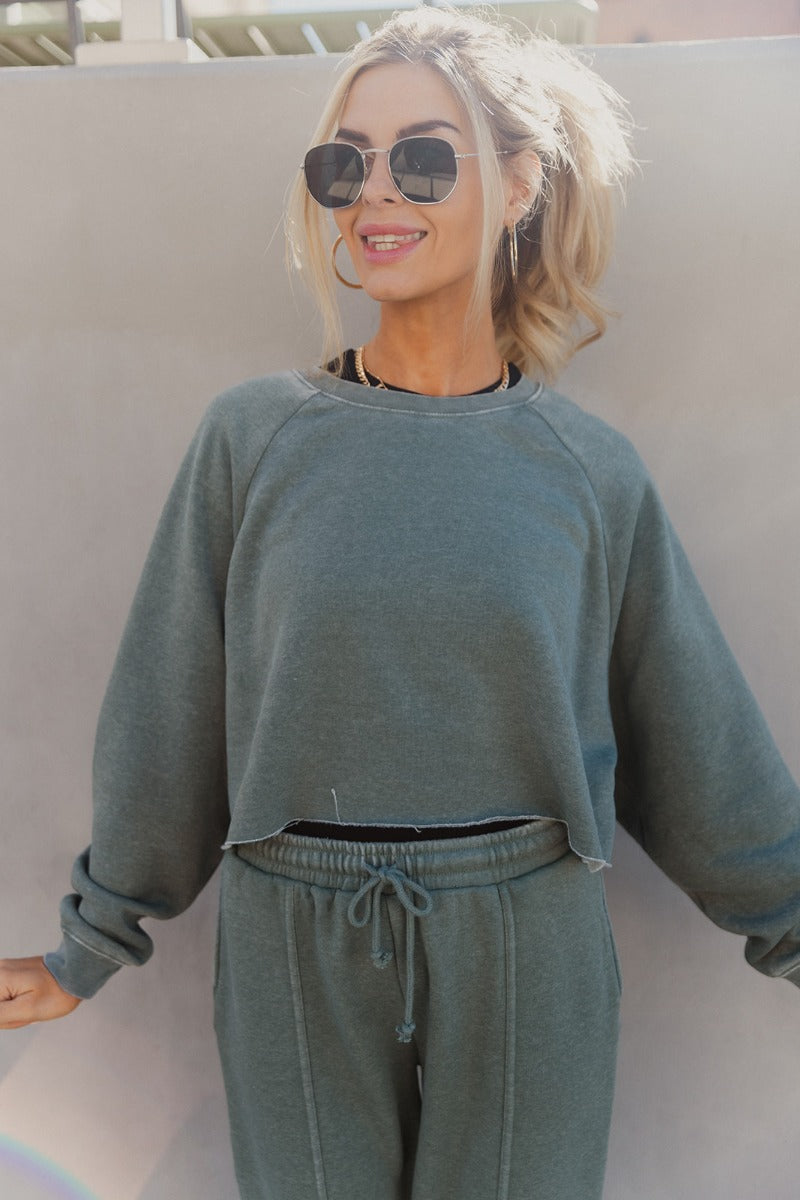 Front view of model wearing the Mia Grey Green Knit Long Sleeve Sweatshirt which features gray green knit fabric, cropped waist, raw hem, round neckline and long sleeves with cuffs.