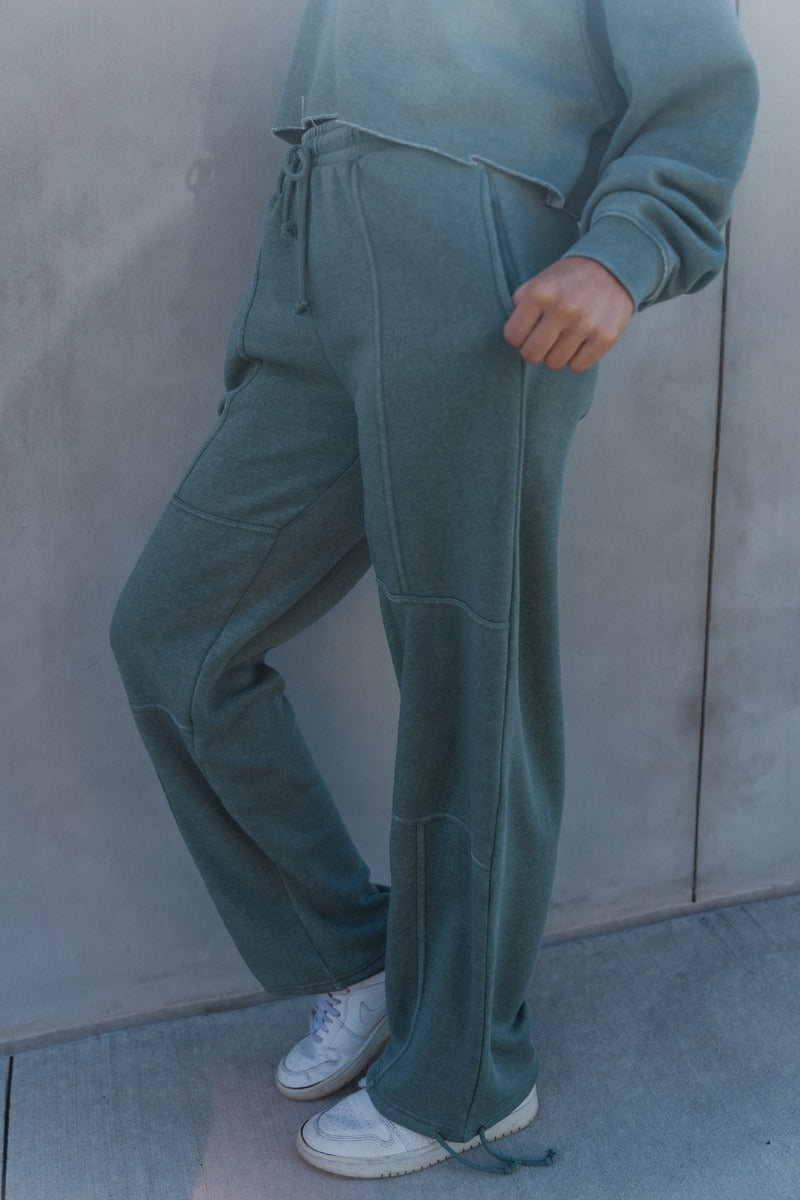 Side view of model wearing the Mia Grey Green Knit Pants which features gray green knit fabric, ribbed hem details, two front pockets, one back pockets, elastic waistband with drawstring ties and wide pant legs with drawstring ties at the ankles.
