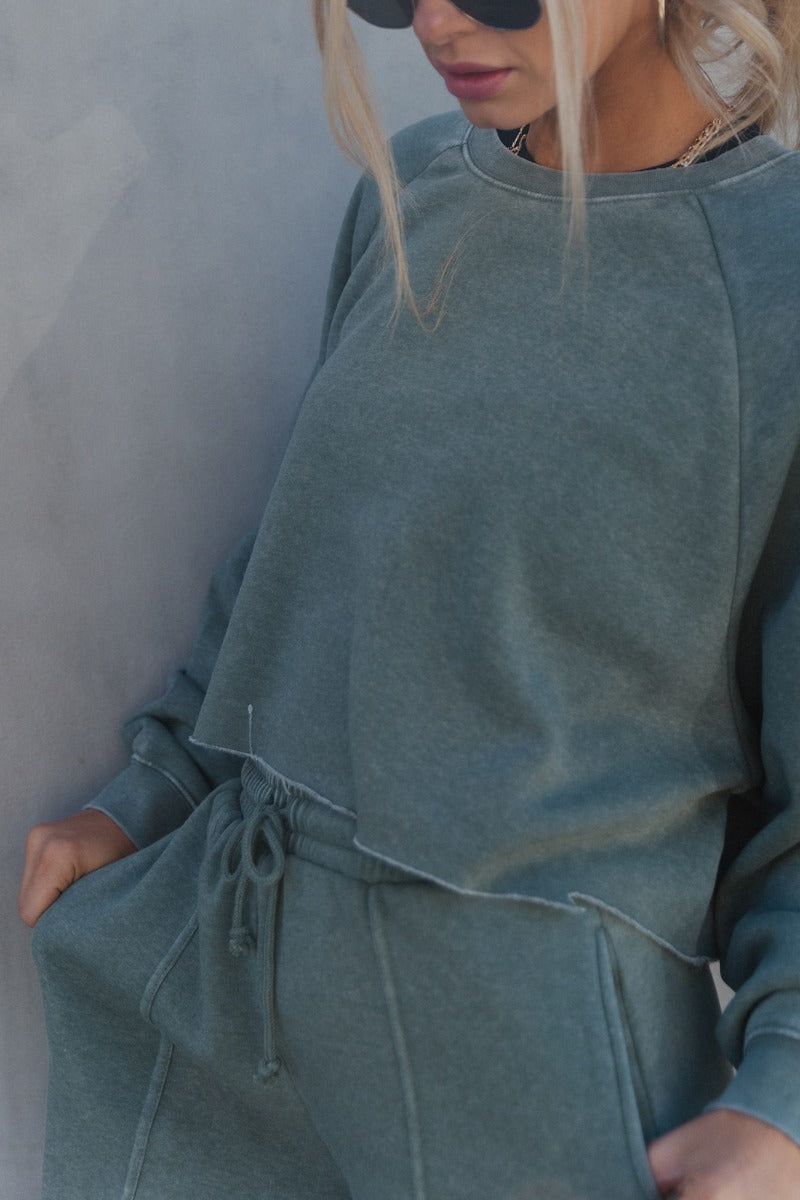 Close up side view of model wearing the Mia Grey Green Knit Long Sleeve Sweatshirt which features gray green knit fabric, cropped waist, raw hem, round neckline and long sleeves with cuffs.