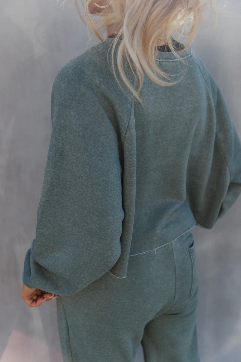 Back view of model wearing the Mia Grey Green Knit Long Sleeve Sweatshirt which features gray green knit fabric, cropped waist, raw hem, round neckline and long sleeves with cuffs.