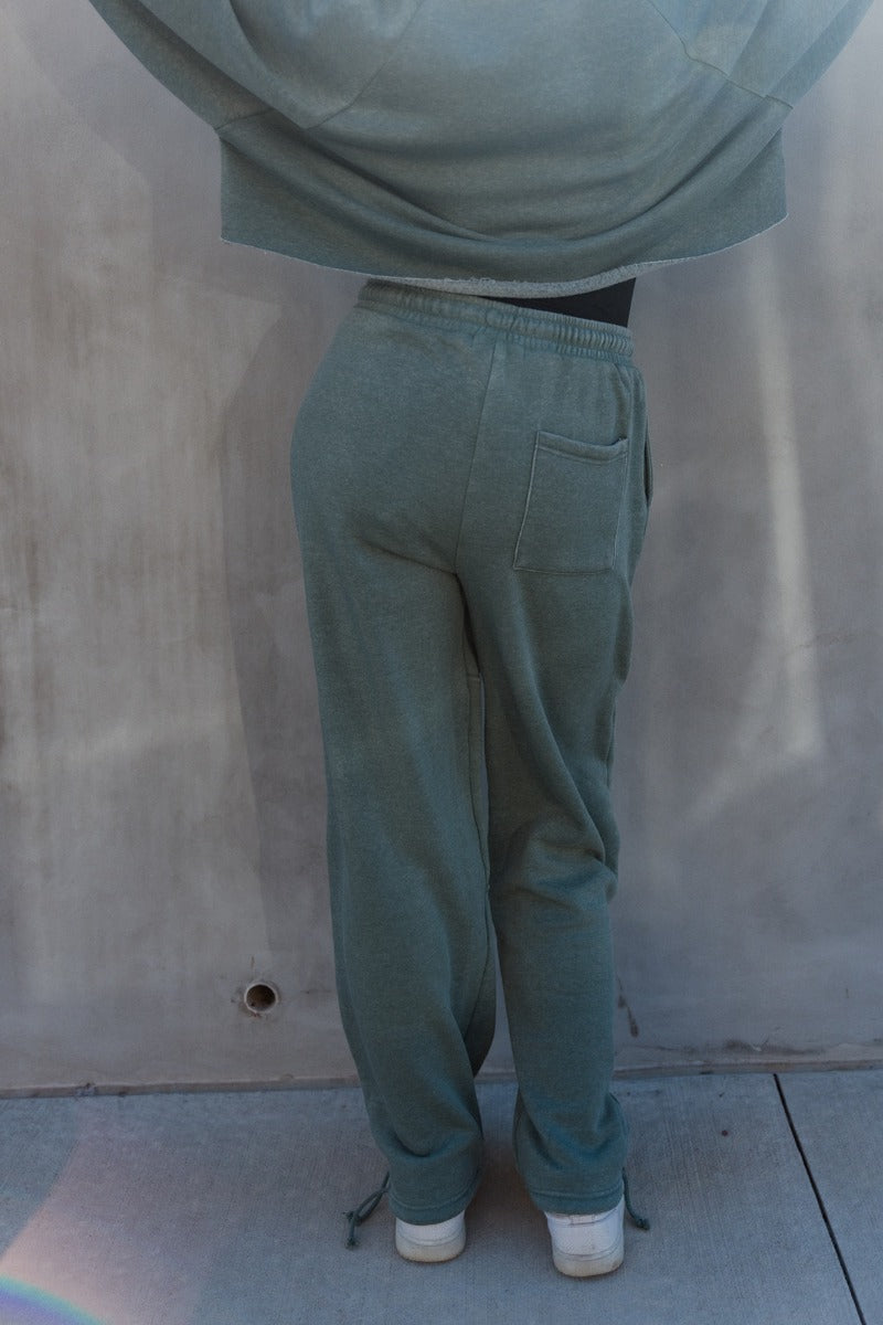 Back view of model wearing the Mia Grey Green Knit Pants which features gray green knit fabric, ribbed hem details, two front pockets, one back pockets, elastic waistband with drawstring ties and wide pant legs with drawstring ties at the ankles.