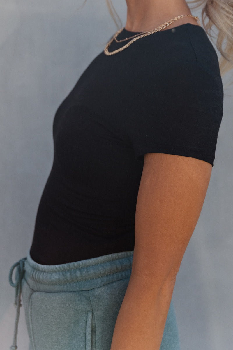 Close up side view of model wearing the Amelia Black Short Sleeve Bodysuit which features black knit fabric, round neckline, short sleeves and thong bottom with button snap closure.