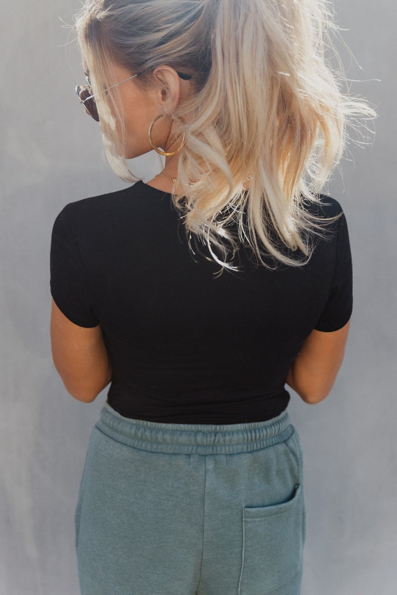 Back view of model wearing the Amelia Black Short Sleeve Bodysuit which features black knit fabric, round neckline, short sleeves and thong bottom with button snap closure.