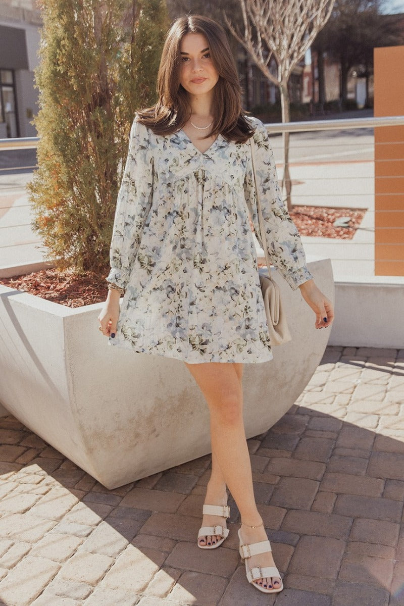 Full body front view of model wearing the Evelyn Blue Multi Floral Mini Dress that has white fabric with a floral pattern, mini length, two covered buttons, a round neckline with a plunge detail, and long sleeves with elastic wrists.