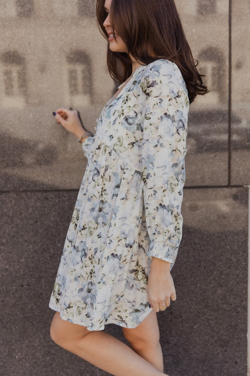 Side view of model wearing the Evelyn Blue Multi Floral Mini Dress that has white fabric with a floral pattern, mini length, two covered buttons, a round neckline with a plunge detail, and long sleeves with elastic wrists.