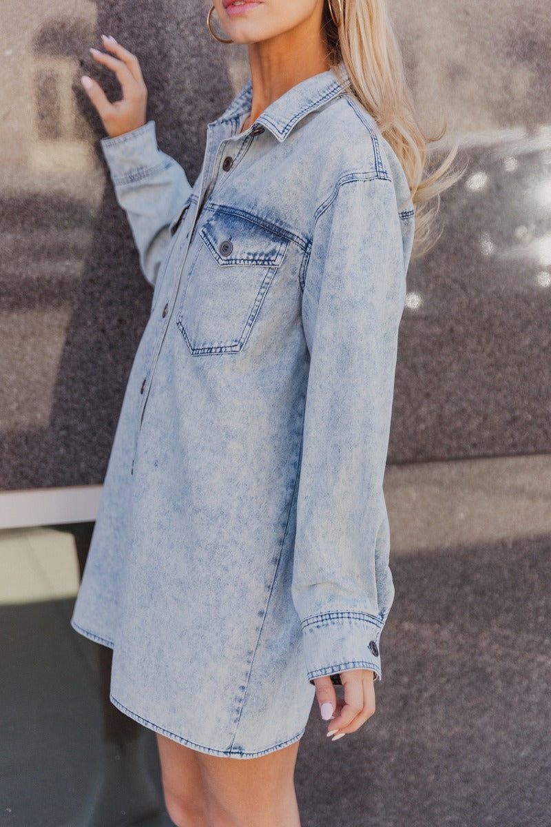 Side view of model wearing the Jasmine Light Denim Wash Buttoned Long Sleeve Dress which features light wash denim fabric, button up closures, mini length, two front chest buttoned pockets, a collared neckline, and long sleeves with buttoned cuffs.