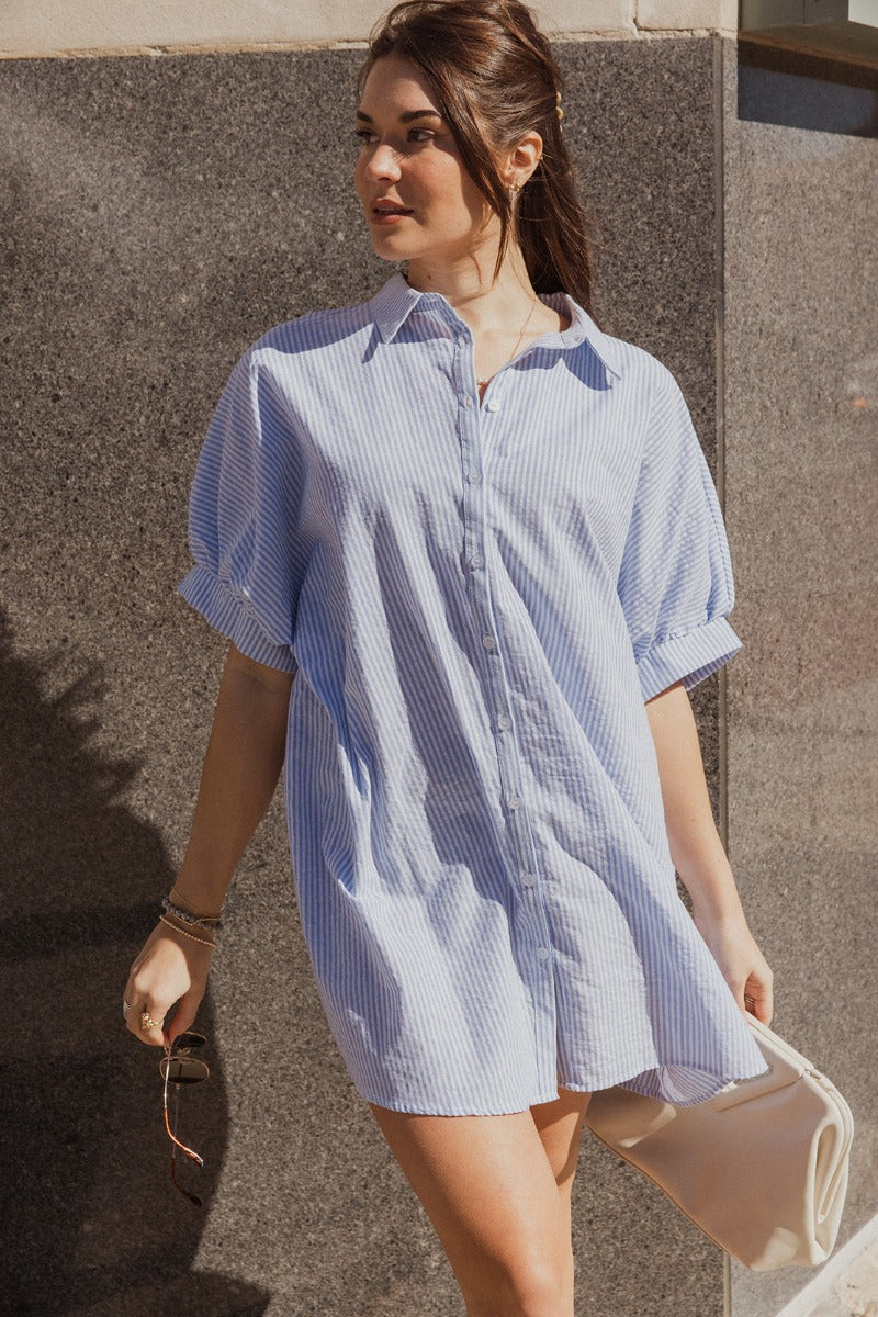front view of model wearing the Alanna Blue & White Striped Button Front Dress that has light blue and white cotton fabric with a striped pattern, white button up front closures, an oversized fit, a collar, and short sleeves.
