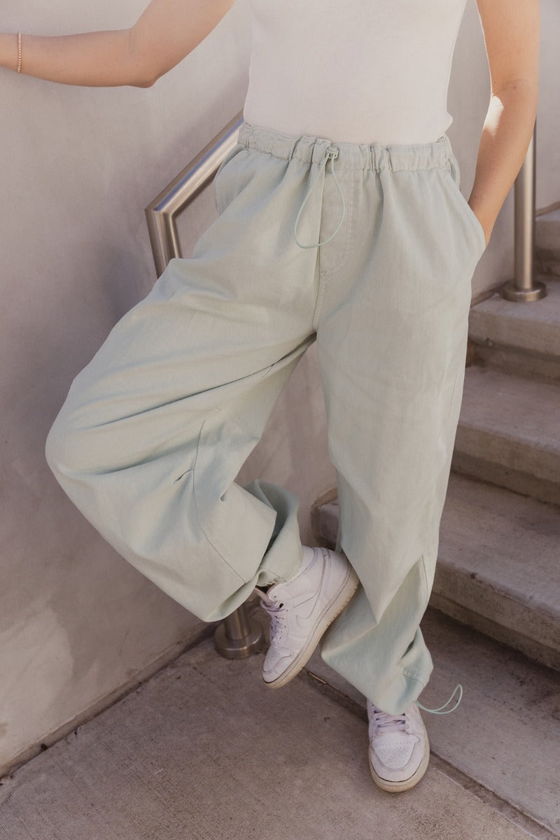 Front view of model wearing the Cecilia Mint Green Pants which features mint denim fabric, two front slit pockets, two back pockets, elastic waistband with drawstring and wide pant legs with drawstrings at the ankles.