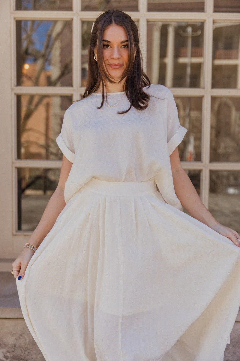 Front view of model wearing the Amelia Off White Midi Skirt which features ivory light weight fabric, midi length, ivory lining, a monochrome pattern design, two slit side pockets, and an elastic waistband.