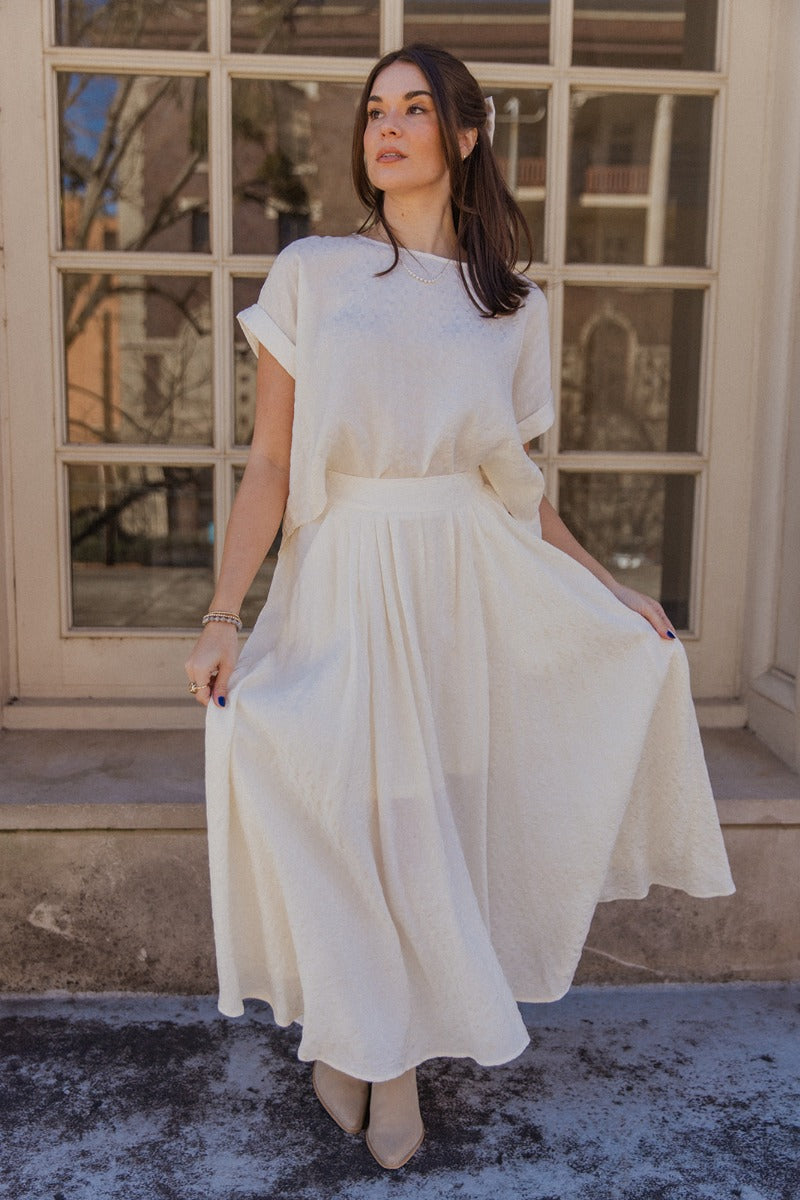 Full body view of model wearing the Amelia Off White Midi Skirt which features ivory light weight fabric, midi length, ivory lining, a monochrome pattern design, two slit side pockets, and an elastic waistband.