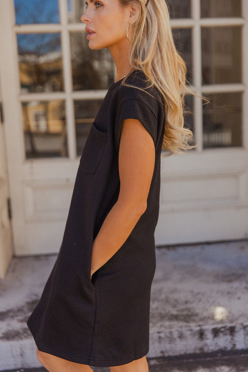Side view of model wearing the Palmer Black Short Sleeve Dress which features light taupe knit fabric with a monochrome block stripe design, mini length, two slit side pockets, a left front chest pocket, a round neckline and sleeveless.