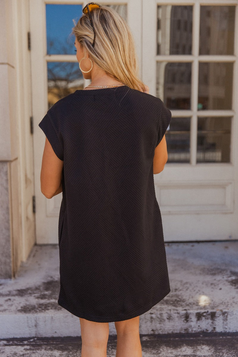 Back view of model wearing the Palmer Black Short Sleeve Dress which features light taupe knit fabric with a monochrome block stripe design, mini length, two slit side pockets, a left front chest pocket, a round neckline and sleeveless.