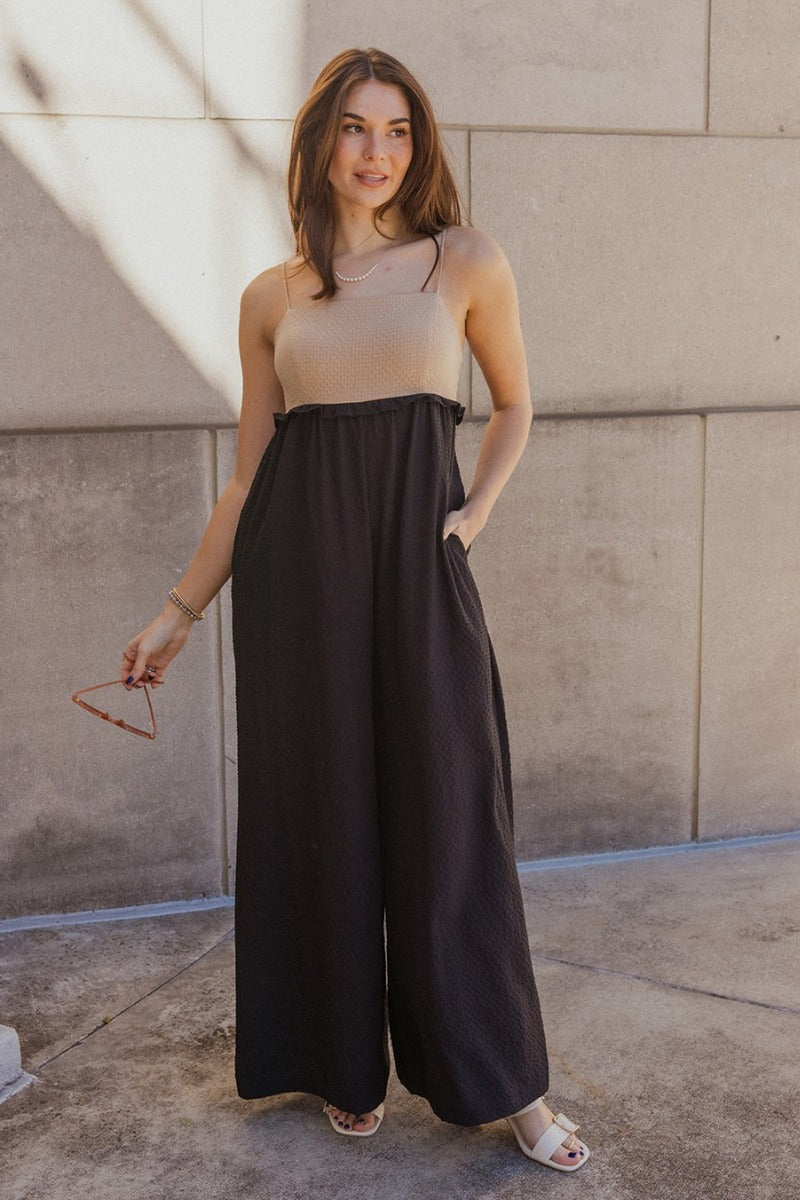 Full body front view of model wearing the Gabriella Black & Taupe Sleeveless Jumpsuit that has black and taupe textured fabric, wide pant legs, two side slit pockets, ruffle details, a square neck, adjustable straps, and a smocked back.