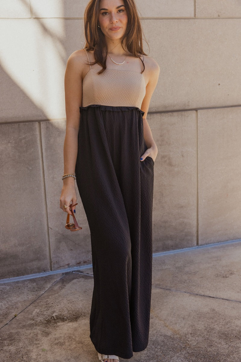 front view of model wearing the Gabriella Black & Taupe Sleeveless Jumpsuit that has black and taupe textured fabric, wide pant legs, two side slit pockets, ruffle details, a square neck, adjustable straps, and a smocked back.