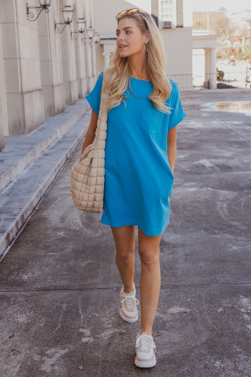 Full body front view of model wearing the Skylar Blue Short Sleeve Mini Dress that has ocean blue cotton fabric, mini length, two side pockets, a left chest pocket, a round neck, and short sleeves with folded hems.